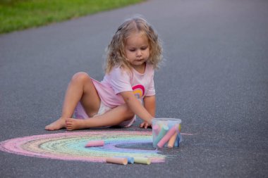 Küçük kız yazın asfalta renkli tebeşirle parlak bir gökkuşağı çiziyor.