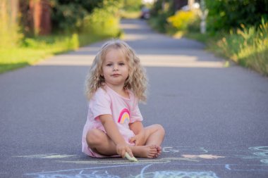 Sevimli küçük kız asfalt yolda tebeşirle renkli bir gökkuşağı çiziyor..