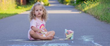 Sevimli küçük kız asfalt yolda tebeşirle renkli bir gökkuşağı çiziyor..