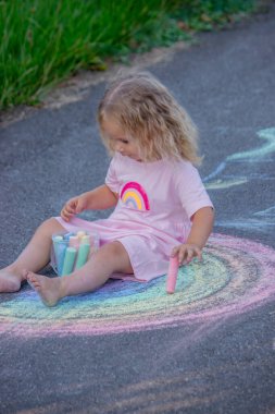 Sevimli küçük kız asfalt yolda tebeşirle renkli bir gökkuşağı çiziyor..