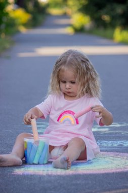 Sevimli küçük kız asfalt yolda tebeşirle renkli bir gökkuşağı çiziyor..