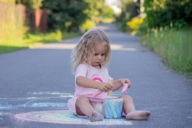 Sevimli küçük kız asfalt yolda tebeşirle renkli bir gökkuşağı çiziyor..