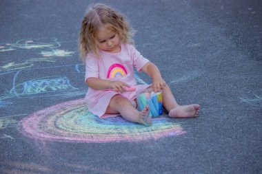 Sevimli küçük kız asfalt yolda tebeşirle renkli bir gökkuşağı çiziyor..