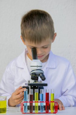 Bilim deneyi sırasında beyaz laboratuar önlüklü küçük çocuk mikroskopla bakıyor..