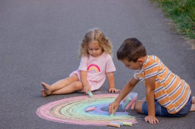 İki mutlu çocuk yaz günü asfalt üzerine tebeşirle renkli gökkuşağı çiziyor..