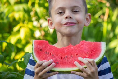 Güneşli bir günde yeşil bir bahçede taze karpuz yiyen küçük bir çocuk.