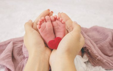 Annenin elleri, pembe tekstil arka planında küçük bebek ayaklarını tutuyor.