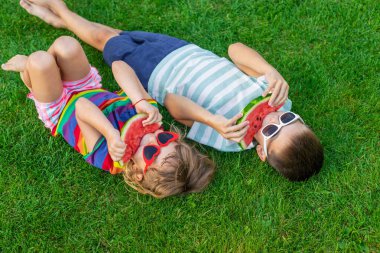 Güneş gözlüklü mutlu çocuklar yeşil çimlerde karpuz dilimleri yiyorlar..