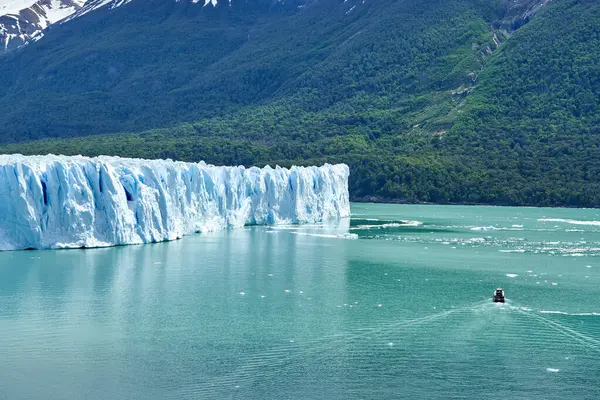 アルゼンチンのパタゴニアにある氷河国立公園にあるペリト モレノ氷河の青い氷 前景のラゴ アルゼンチノのターコイズブルーの水に観光船で 暗い緑の森と遠くのアンデス山脈の覆われた山々を蒔く — ストック写真