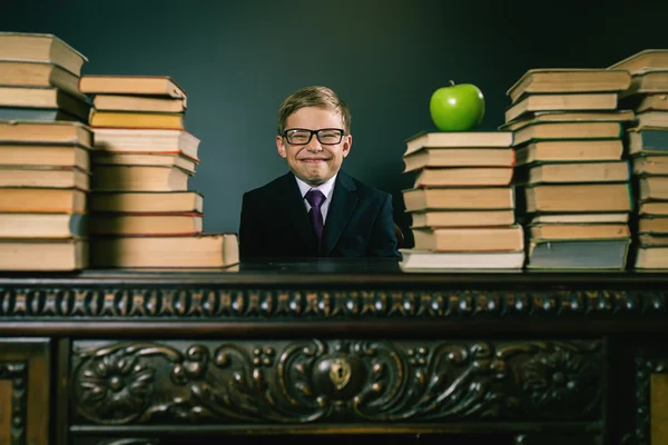 Chico de escuela astuto sentado a la mesa con muchos libros — Foto de Stock