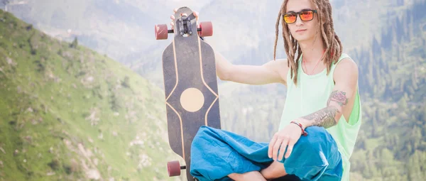 Hippie giovane e bello uomo con longboard skateboard in montagna — Foto Stock