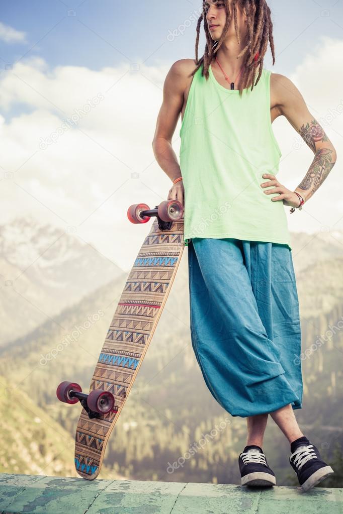 Hippie young and handsome man with longboard skateboard at mountain