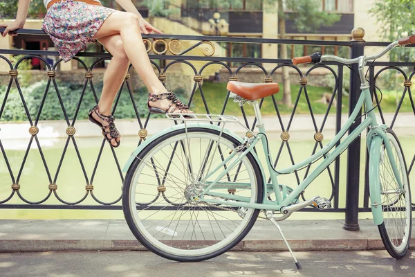 Chica joven sentada en la valla cerca de la bicicleta vintage en el parque — Foto de Stock