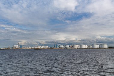 Büyük beyaz petrol depolama tankları ve bulutlu bir gökyüzünün altında rıhtım yakınında mavi vinçleri olan sanayi limanı. Deniz kenarındaki petrol depolama ve lojistik tesisi.