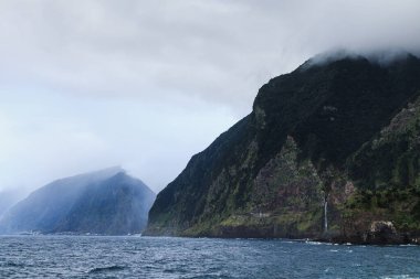 Okyanus kıyısındaki kayalıklar. Yüksek dağların bulutlara değdiği güzel bir manzara, uçurumdan okyanusa düşen bir şelale..