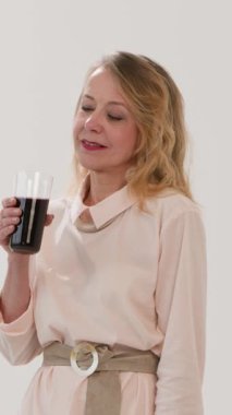 Olgun bir kadın nar suyu içer ve elleri oda lambasında olan bir kalp gösterir. Gülümseyen sarışın bayan stüdyo dikey videosunda taze sıkılmış içecekten hoşlanıyor. Diyet