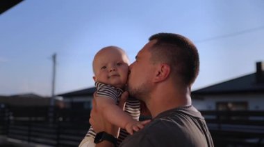 Mutlu genç baba kollarını tutuyor ve kulübedeki küçük çocuğu öpüyor. Yavaş çekimde. Sakallı adam, kırsal kesimde havadaki sevimli oğlunu yetiştirme oyunu oynuyor. Babalığın mutlu anları