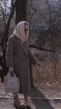 Dikey video. Bahar parkında başörtülü kalın palto giyen yaşlı bir kadın köpekle oyun oynuyor. Aktif kıdemli bayan, güneşli şehir bahçesinde yürüyüş sırasında dost canlısı evcil hayvanla tanışır. Yaşlılıkta sağlıklı bir hayat