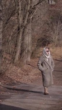 Dikey video. Kıdemli bayan yün başörtülü kalın bir palto giyiyor ve yol boyunca çıplak ağaçların arasında yürüyor. Sakin yaşlı kadın güneşli sonbahar bahçesinde geziniyor. Emekliliğin yaşam tarzı
