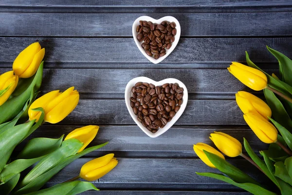 Na drewnianym szarym tle są żółte tulipany i spodki z ziarnem kawy w kształcie ósemki Koncepcja świątecznej wiosennej kartki — Zdjęcie stockowe