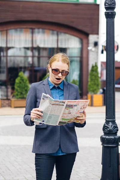 Business blonde in a gray jacket and blue shirt in surprise reads a newspaper on the street