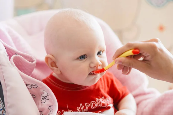 breastfed blonde baby in a red t-shirt tries first complementary foods from a small red spoon on a pink background