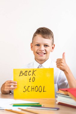 Beyaz gömlekli mutlu sarışın okul çocuğu ders kitapları ve kalemlerin arka planında oturuyor ve okula sarı bir tabela asıyor ve baş parmak işareti gösteriyor.
