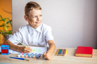 Beyaz gömlekli sarışın bir çocuk, bir albümde arka planda boya, fırça, kalem ve kırmızı bir kitapla birlikte resim çiziyor, okulda bir masada oturuyor.
