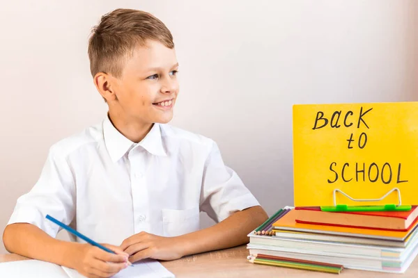 白いシャツを着た幸せなブロンドの学童は 教科書や鉛筆の背景を背景に机に座っている黄色の看板を学校に戻す — ストック写真