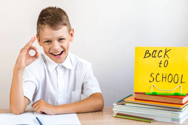 白いシャツを着た幸せなブロンドの女子高生は 教科書や鉛筆の背景を背景に机に座って黄色の看板をバック学校に示し Okジェスチャー — ストック写真