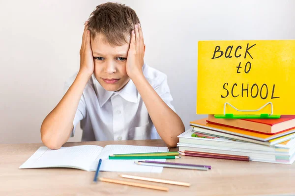 白いシャツを着た金髪の男子生徒が 教科書や鉛筆を背景に机に座り 黄色の看板を掲げて学校に戻ります — ストック写真
