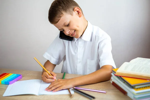 白いシャツを着た感情的な金髪の少年が電話で話していて 机に座っている間にノートに何かを書いています インターネット通信 子供のためのレジャー活動 — ストック写真