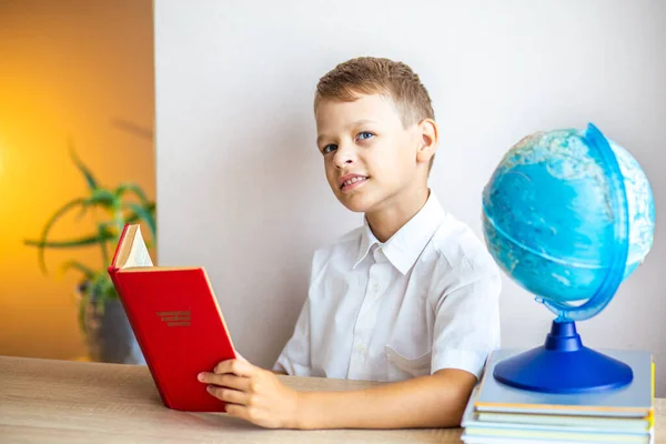 a blond boy in a white shirt reads a book in a red cover on the background of the globe, sitting at the table