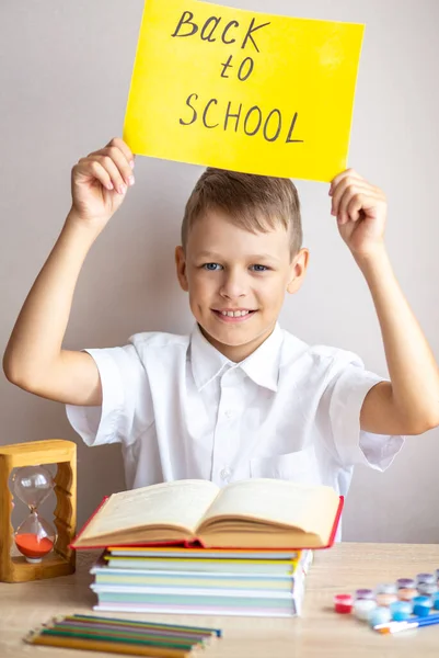 穿着白衬衫的快乐的金发碧眼的男生坐在书桌前 书桌后面是课本和铅笔 上面有一个黄色的标志 然后返回学校 — 图库照片