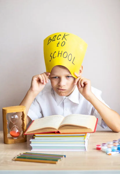 白いシャツを着た金髪の男子生徒が 教科書や鉛筆を背景に机に座り 黄色の看板を掲げて学校に戻ります — ストック写真