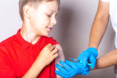 Beyaz şapkalı, tek kullanımlık maskeli ve mavi eldivenli bir hemşire aşıyı kırmızı tişörtlü cesur bir çocuğun omzuna şırıngayla enjekte ediyor..