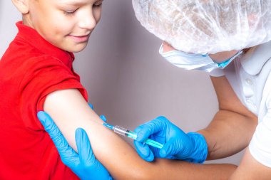 Beyaz şapkalı, tek kullanımlık maskeli ve mavi eldivenli bir hemşire aşıyı kırmızı tişörtlü cesur bir çocuğun omzuna şırıngayla enjekte ediyor..