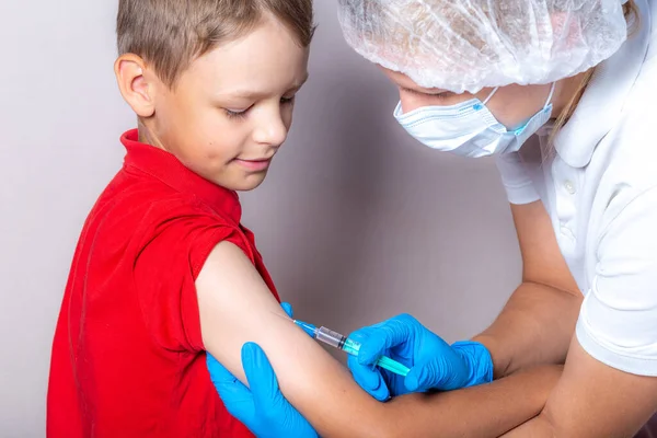 A nurse in a white cap, disposable mask and blue gloves injects the vaccine through a syringe into the shoulder of a brave boy in a red T-shirt.