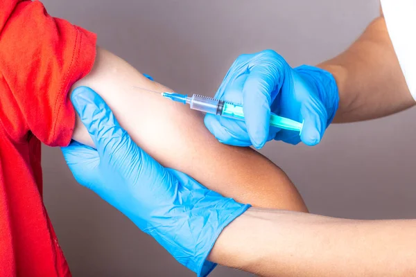 A nurse in a white cap, disposable mask and blue gloves injects the vaccine through a syringe into the shoulder of a brave boy in a red T-shirt.