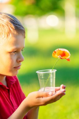 Kırmızı tişörtlü sarışın çocuk bir bardak su ve temiz havada bir çiçek tutuyor. Ekolojik bir sorun. Doğayı kurtar.