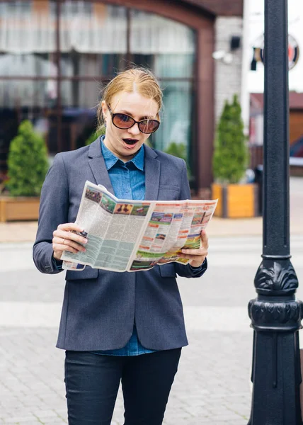 a blonde girl in a gray jacket and brown glasses is reading the fresh press in surprise on the street on a bright day