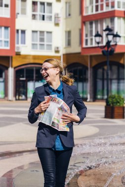 Gri ceketli ve gözlüklü sarışın kız zıplıyor ve elinde bir gazeteyle sokakta eğleniyor..