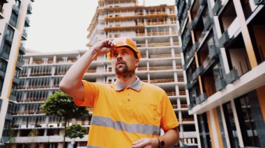 Turuncu üniformalı sakallı işçi inşaat sırasında binanın dışında dikilirken kafasına kask takıyor. İş kıyafetleri içinde beyaz erkek inşaat işçisi koruyucu bir kask takıyor..