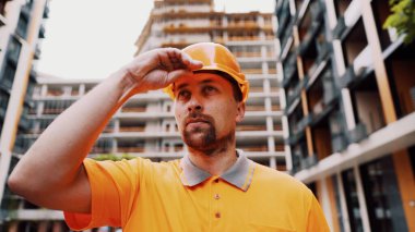 Ciddi bir Kafkasyalı inşaat alanında duruyor ve turuncu bir şapka takıyor. Öncelikle güvenlik, inşaat mesleği. Düşük açılı çekim. Foreman inşaat sürecini inceleyip, miğfer takıyor..