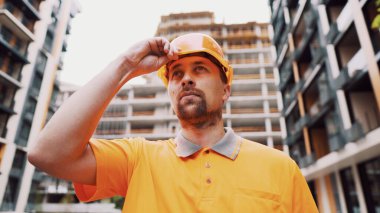 İnşaat işçisi, şantiyede kaskını takıyor. Önce güvenlik, meslek anlayışı. İnşaatçı sert şapka üniformasını giyer ve işe hazırlanır. Adam turuncu kapaklı şantiye yapıyor..