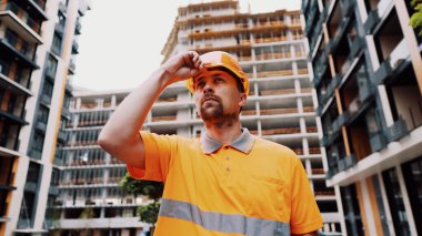 Ciddi bir Kafkasyalı inşaat alanında duruyor ve turuncu bir şapka takıyor. Öncelikle güvenlik, inşaat mesleği. Düşük açılı çekim. Foreman inşaat sürecini inceleyip, miğfer takıyor..