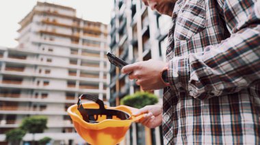 Caucasian male construction worker in protective helmet use smartphone near building that is being built. Engineer checks construction process, makes notes on phone. Foreman chatting in application. clipart