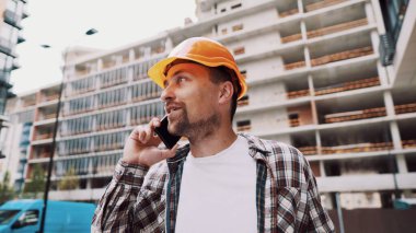 Turuncu kasketli ve ekose gömlekli beyaz erkek inşaat işçisi şantiyede telefonla konuşuyor. Mimarlık teması. Erkek işi. Foreman inşaat sürecini akıllı telefonla kontrol ediyor.