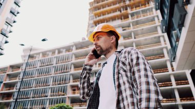 Turuncu kasketli ve ekose gömlekli beyaz erkek inşaat işçisi şantiyede telefonla konuşuyor. Mimarlık teması. Erkek işi. Foreman inşaat sürecini akıllı telefonla kontrol ediyor.