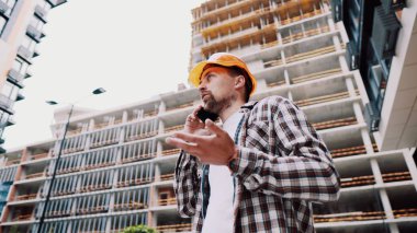 İnşaat alanında duran genç işçinin portresi. İnşaat alanında mühendis telefonda konuşuyor. Kask kontrolündeki yapıcı cep telefonuna göre planlandığı gibi.